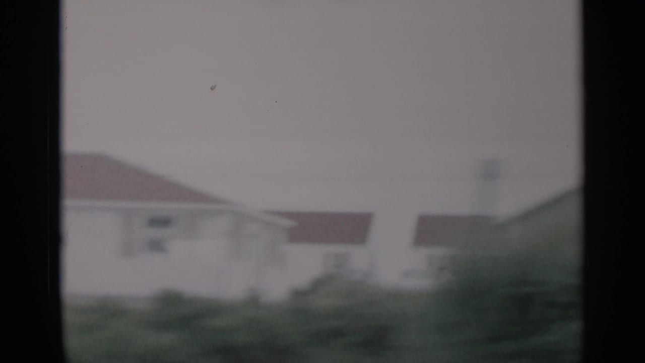 1956 Cape Cod home-movie frame showing a small residence and roadside grass through a dark vehicle window frame.