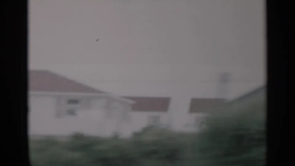 1956 Cape Cod home-movie frame showing a small residence and roadside grass through a dark vehicle window frame.