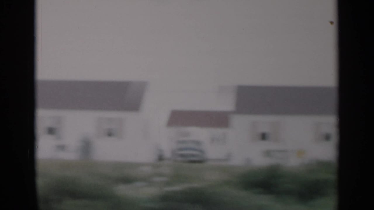 Cape Cod residential drive-by with light house exteriors, rooflines, grass, and a parked vehicle visible in soft 8mm focus.