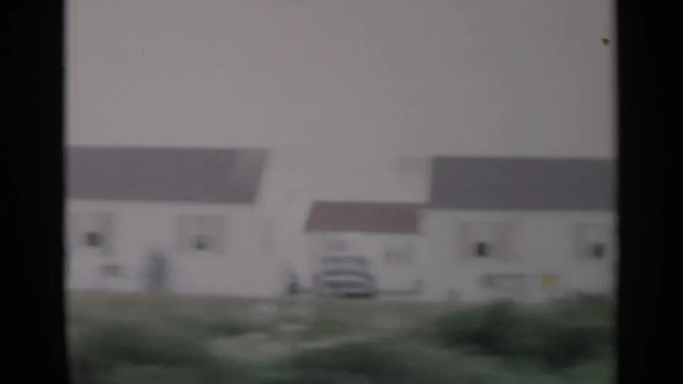 Cape Cod residential drive-by with light house exteriors, rooflines, grass, and a parked vehicle visible in soft 8mm focus.
