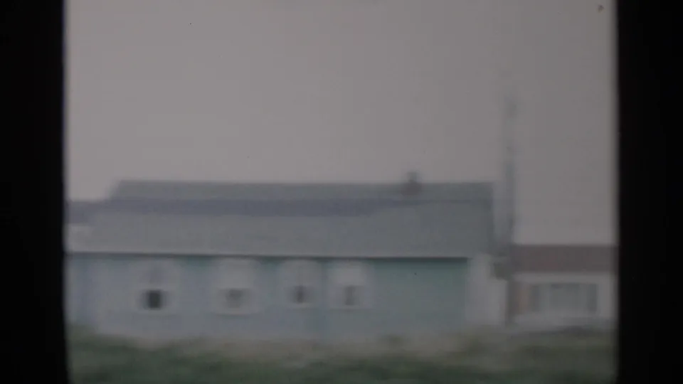 1956 home-movie frame with a soft view of a Cape Cod house exterior, low roofline, grass, and a utility pole.
