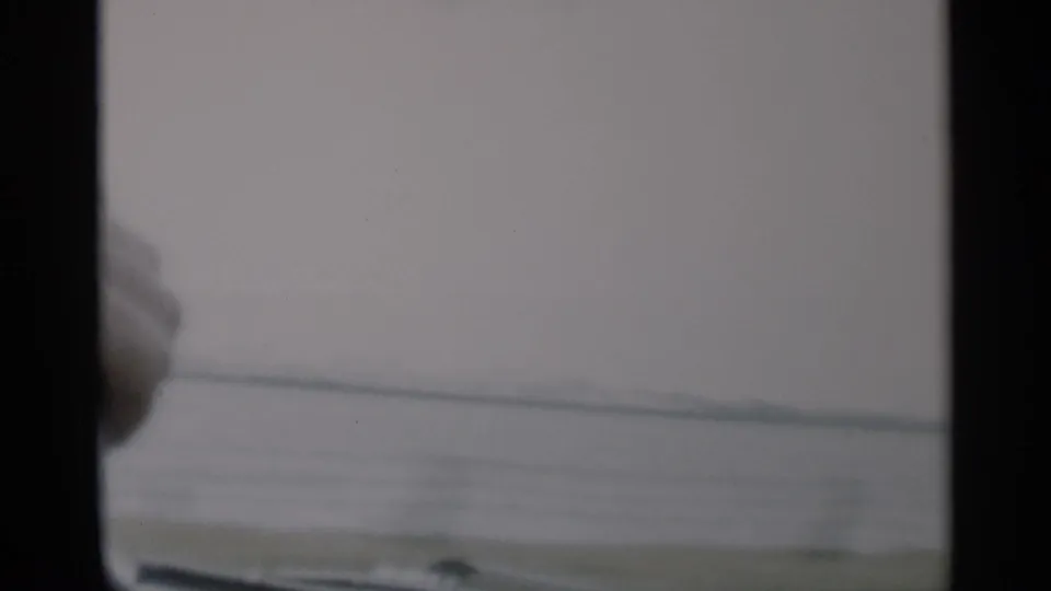 1956 Cape Cod home-movie frame from inside a moving car, looking past windshield posts toward muted water and low shoreline.