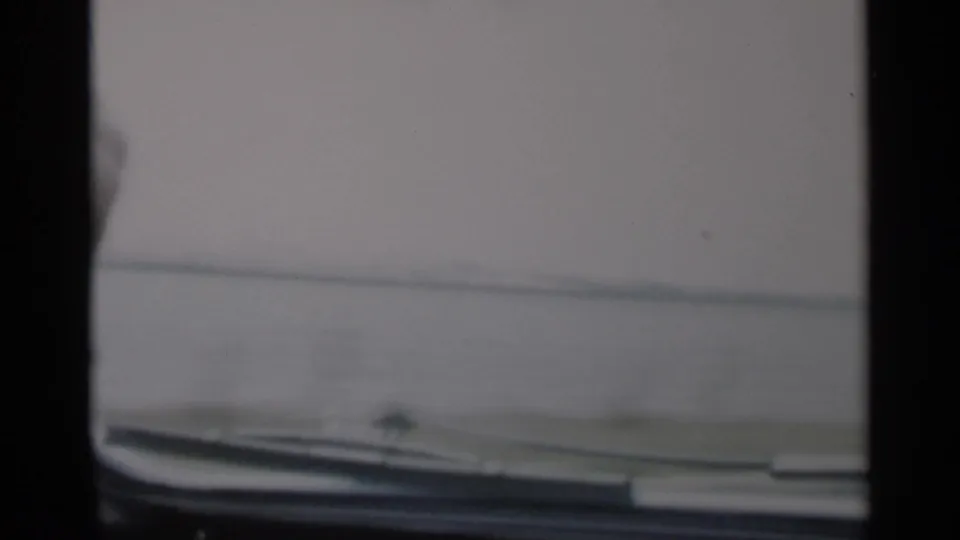 Cape Cod driving view through a mid-century car windshield, with dashboard edge, water, and a hazy shoreline visible.