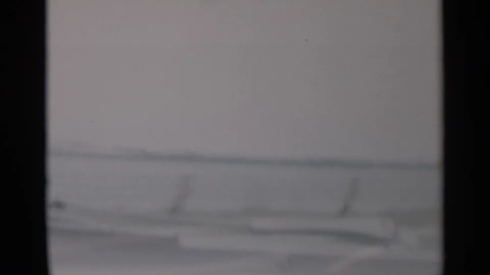 Final 1956 Cape Cod frame with blurred windshield framing, pale coastal water, and a faint shoreline seen from the car.