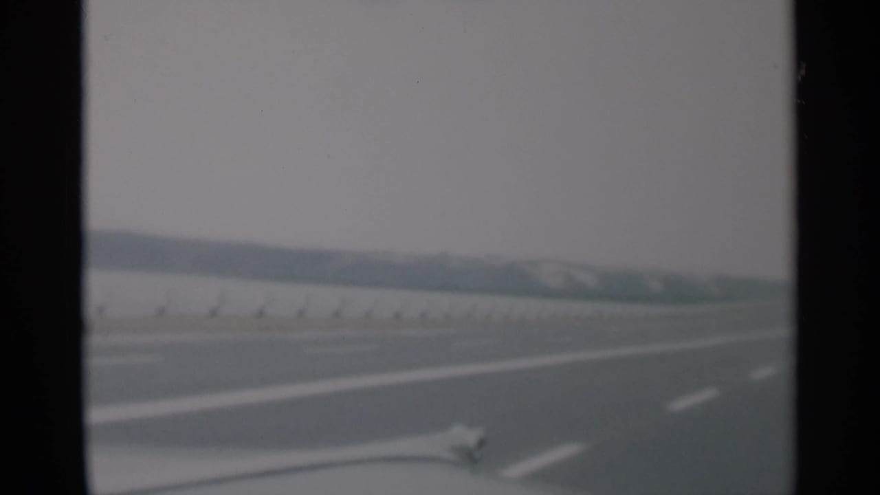 Soft 8mm view through a windshield of an open highway with lane markings and a low barrier.