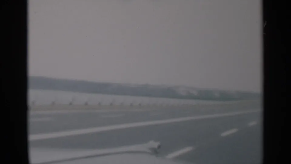 1956 Cape Cod home-movie frame showing a quiet highway from a moving car, with white lane markings, roadside barrier, and hazy horizon.