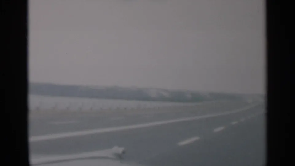 1956 Cape Cod 8mm frame of an empty-looking highway viewed from a car, with muted color, lane markings, and low distant land.
