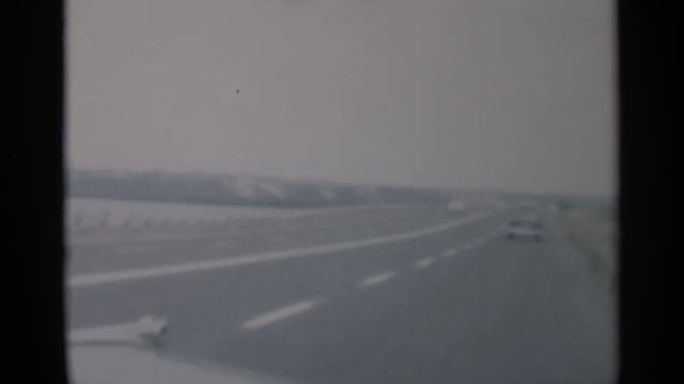 Cape Cod 1956 home-movie frame showing a roadway from a moving car, with a small distant vehicle, lane markings, and muted 8mm color.
