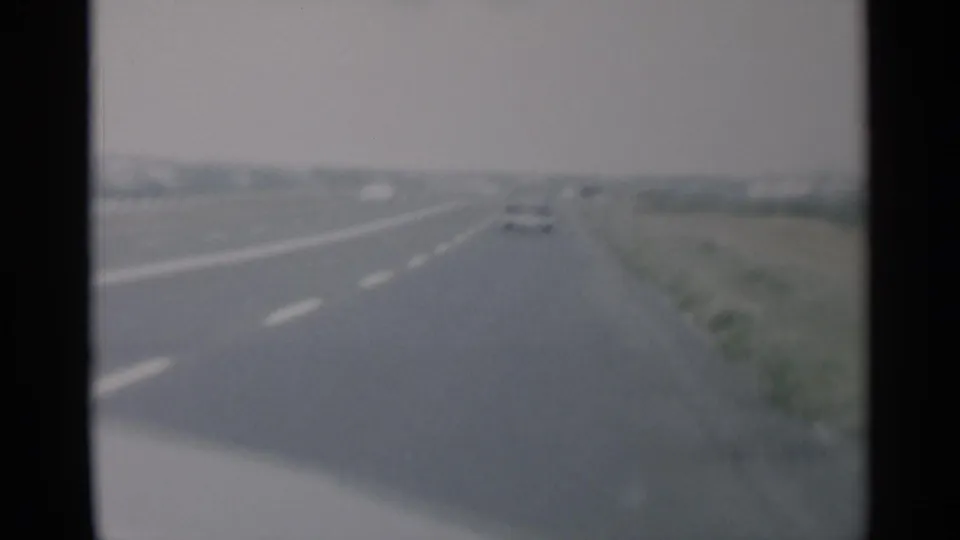 1956 Cape Cod road-travel frame showing cars ahead on a straight highway, with white lane markings, grassy shoulder, and hazy sky.
