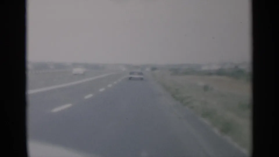 1956 Cape Cod road-trip footage shows a soft forward view down a divided highway with cars ahead and flat roadside land.