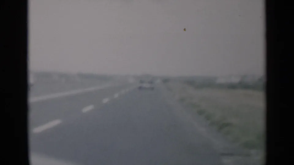 A soft 8mm frame shows the road trip continuing along a broad Cape Cod highway, with one car visible ahead in the distance.