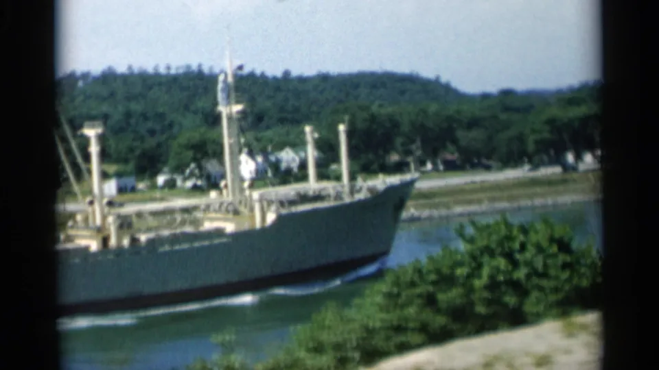 Cape Cod footage shows a large cargo ship moving along a narrow waterway, backed by trees, shoreline homes, and low hills.