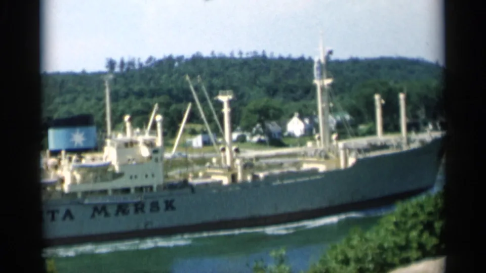 The cargo ship RITA MERSK is visible in the waterway, with its name on the hull, a marked funnel, trees, and coastal homes behind it.