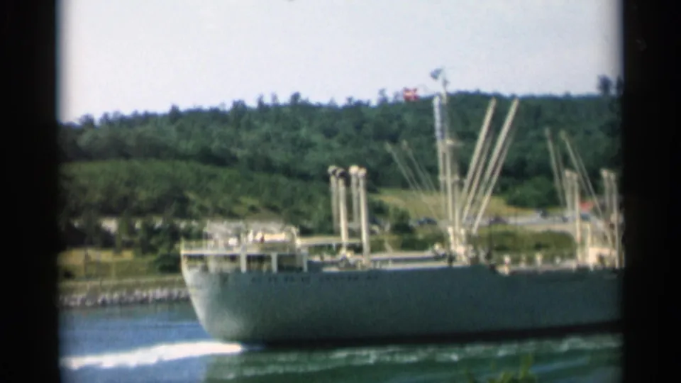 A home-movie frame shows the forward section of the cargo ship entering view, with cranes, water wake, and green bank behind it.