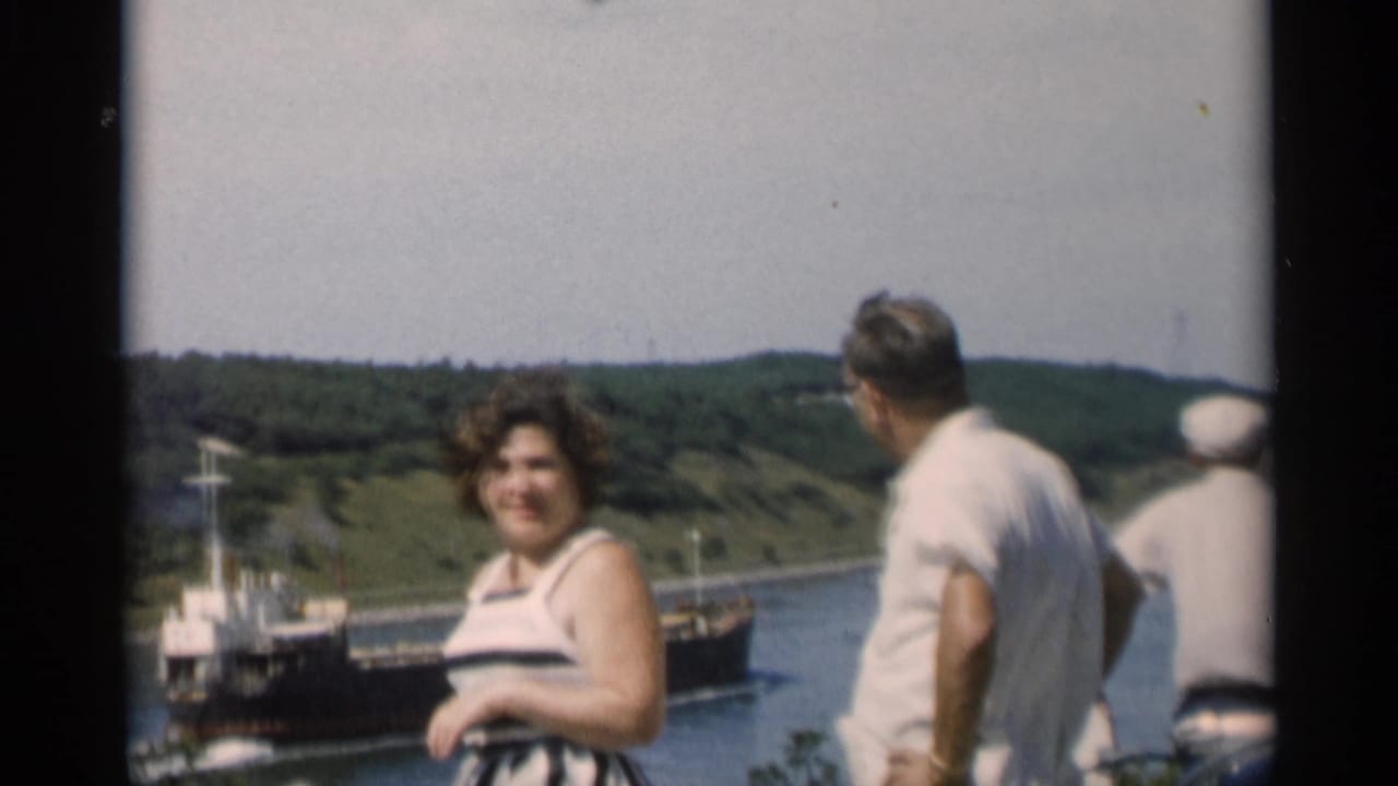 In 1956 Cape Cod home-movie footage, adults stand by the water while a dark ship moves past wooded hills.