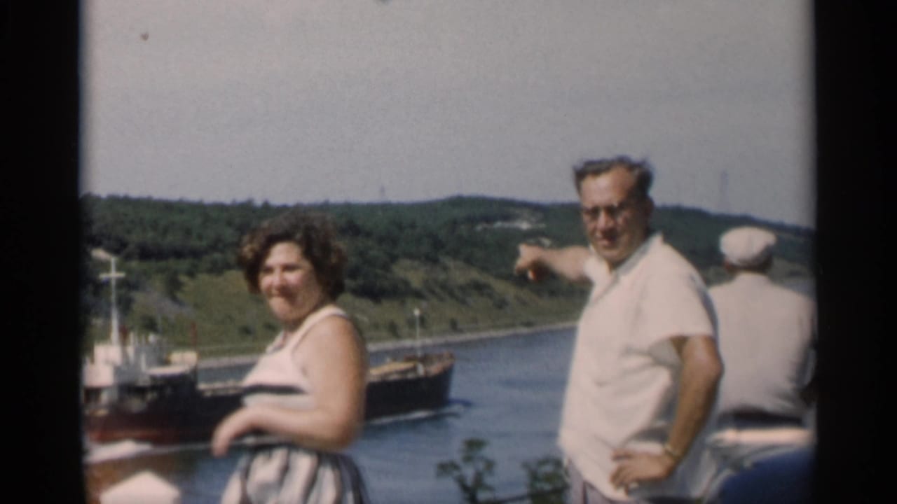 A man in a white shirt points toward a passing ship as a woman in a striped dress stands near Cape Cod water.