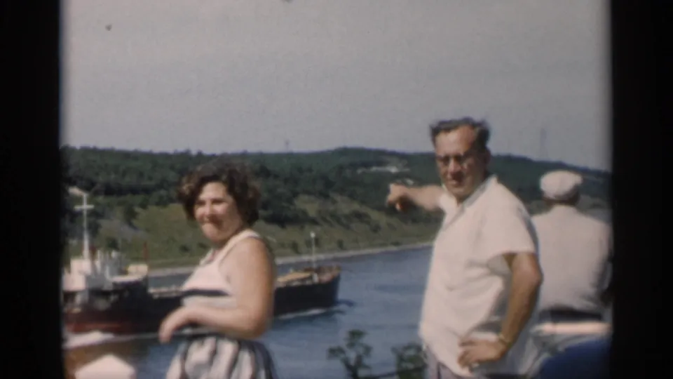 A man in a white shirt points toward a passing ship as a woman in a striped dress stands near Cape Cod water.