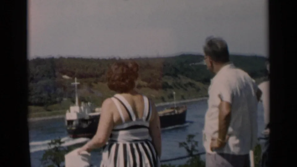 From behind, the adults look out over the Cape Cod waterway toward a dark ship and green shoreline.