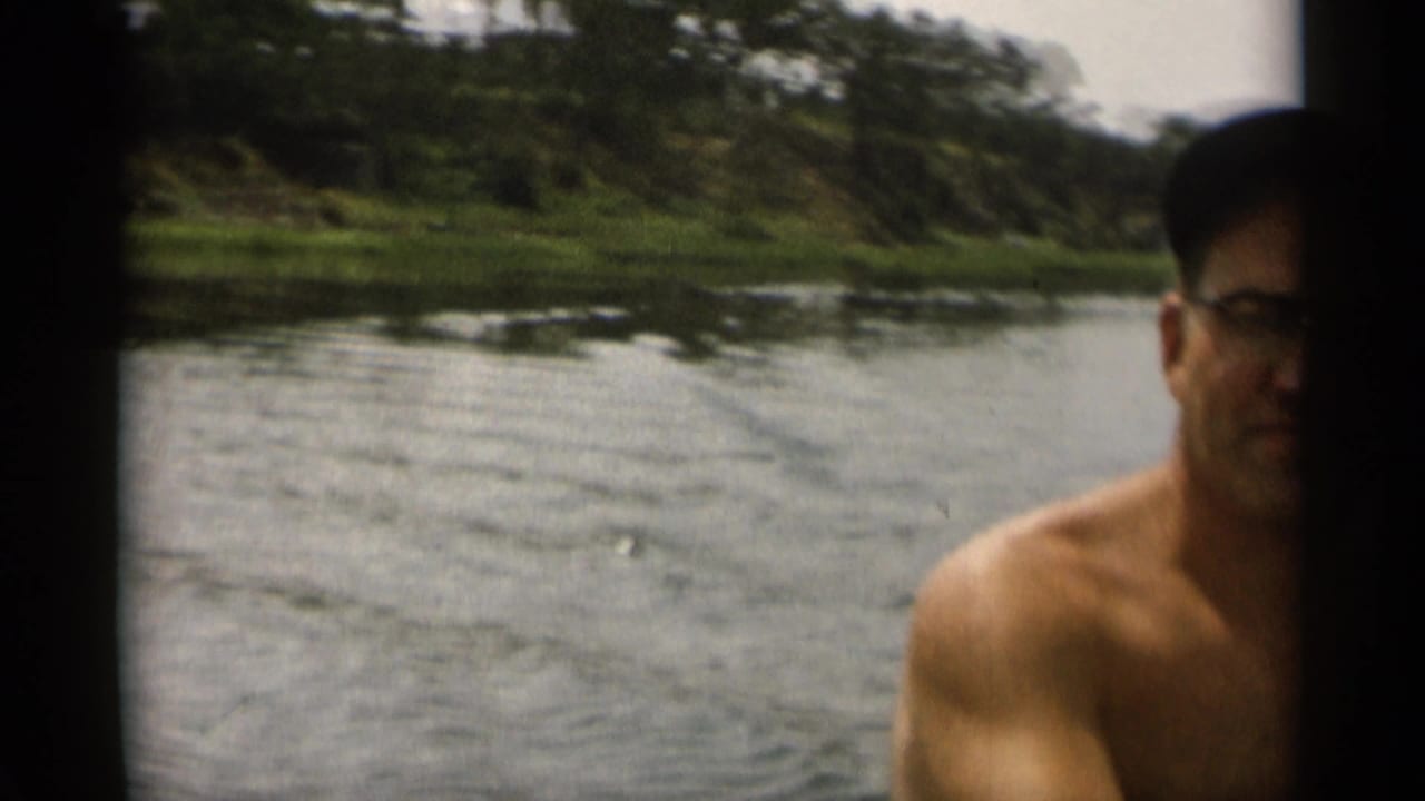 In supported 1956 Cape Cod home-movie footage, a shirtless man with glasses looks toward the camera from the water near a tree-lined shore.
