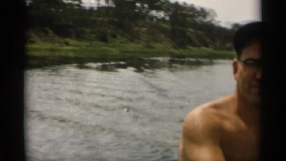 In supported 1956 Cape Cod home-movie footage, a shirtless man with glasses looks toward the camera from the water near a tree-lined shore.