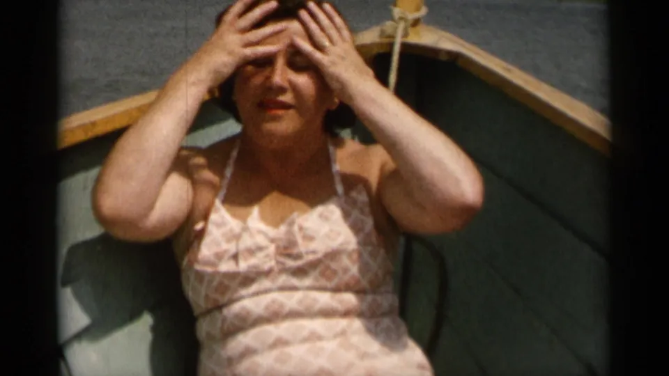 A woman in a patterned swimsuit sits in the wooden boat, raising both hands to adjust her hair under bright sun.