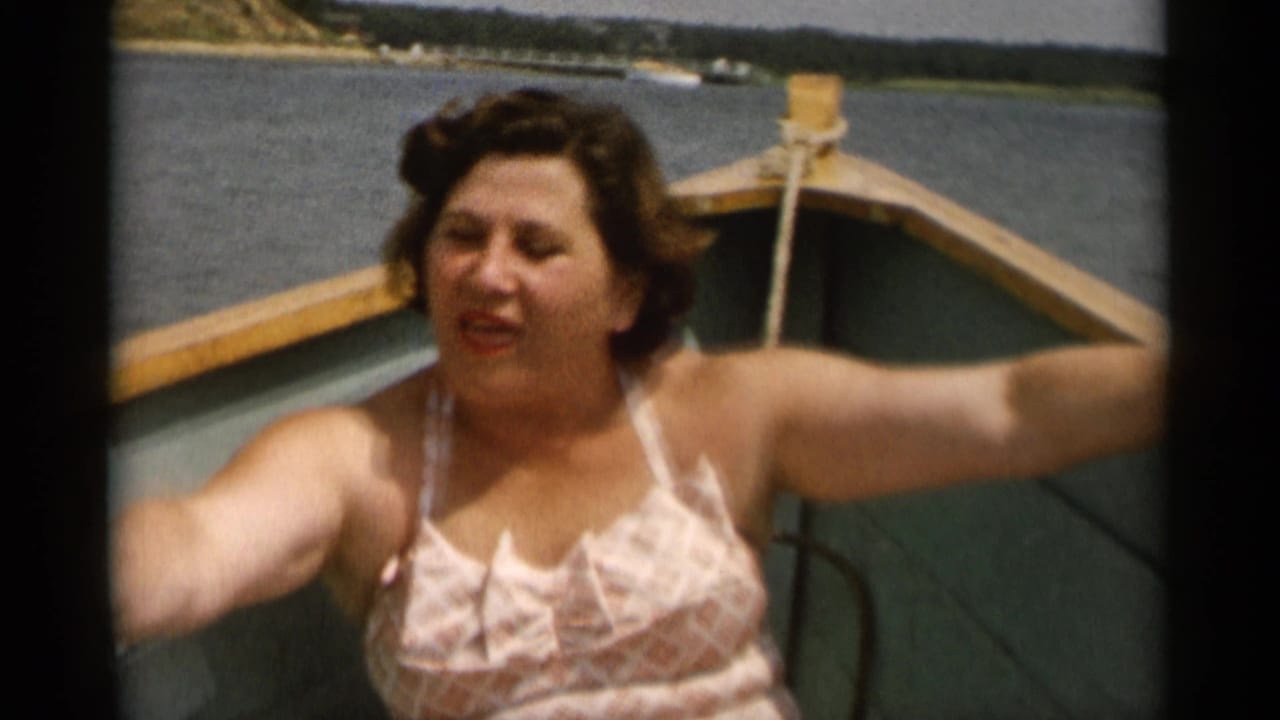 In Cape Cod home-movie footage from 1956, a woman in a patterned swimsuit sits near the bow of a small wooden boat on calm water.