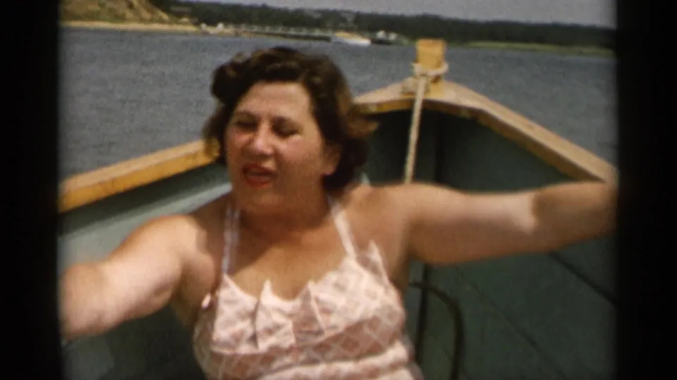 In Cape Cod home-movie footage from 1956, a woman in a patterned swimsuit sits near the bow of a small wooden boat on calm water.
