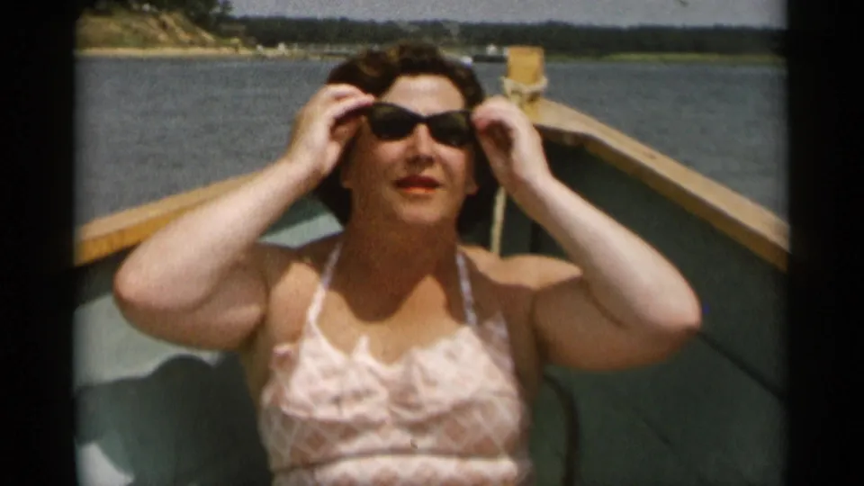 The woman faces the camera and adjusts dark sunglasses while sitting in the boat on Cape Cod water in 1956 home-movie footage.