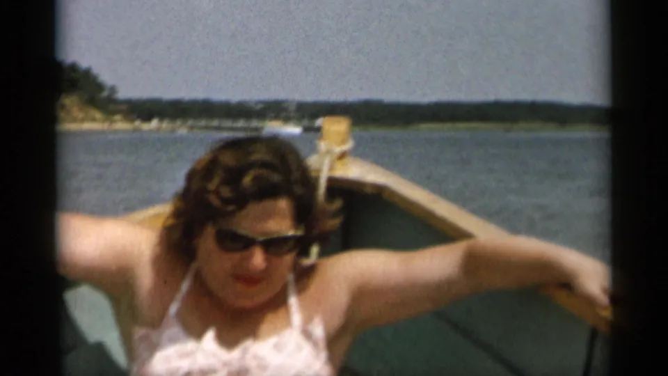 The woman, now wearing sunglasses, sits low in the small boat with calm water and a distant Cape Cod shoreline behind her.
