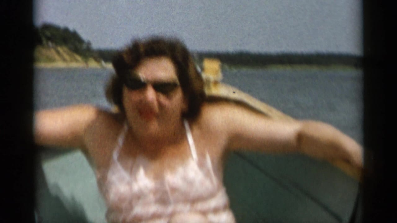 In 1956 Cape Cod footage, a woman in dark sunglasses and a pink patterned swimsuit leans toward the camera from a small boat.