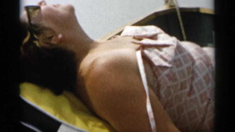 The 1956 home-movie frame shows the woman reclining on a yellow pillow in the boat, her sunglasses and swimsuit visible in close view.