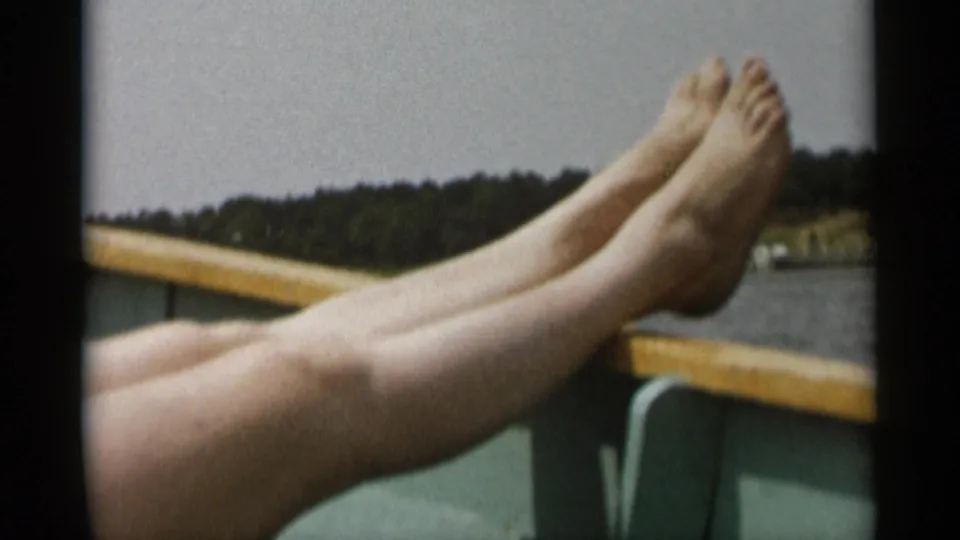 Bare legs and feet rest near a boat rail in 1956 Cape Cod footage, with water and wooded shore in the background.