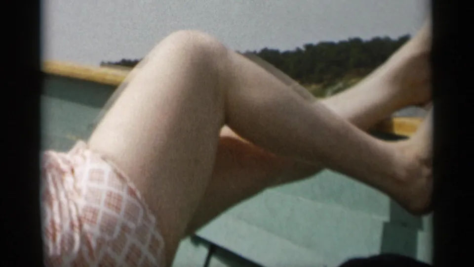 1956 Cape Cod frame of a reclining woman’s bent legs and light patterned dress on a boat near a wooded shoreline.
