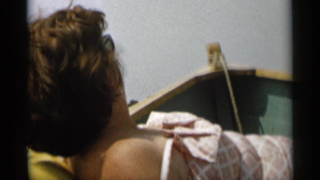 1956 Cape Cod frame of a woman with short hair and sunglasses reclining in a small boat, with a folded towel near the bow.