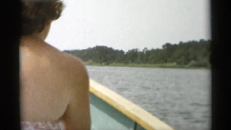 1956 Cape Cod home-movie view from a small boat, with a woman in the foreground looking across lake water toward a tree-lined shore.