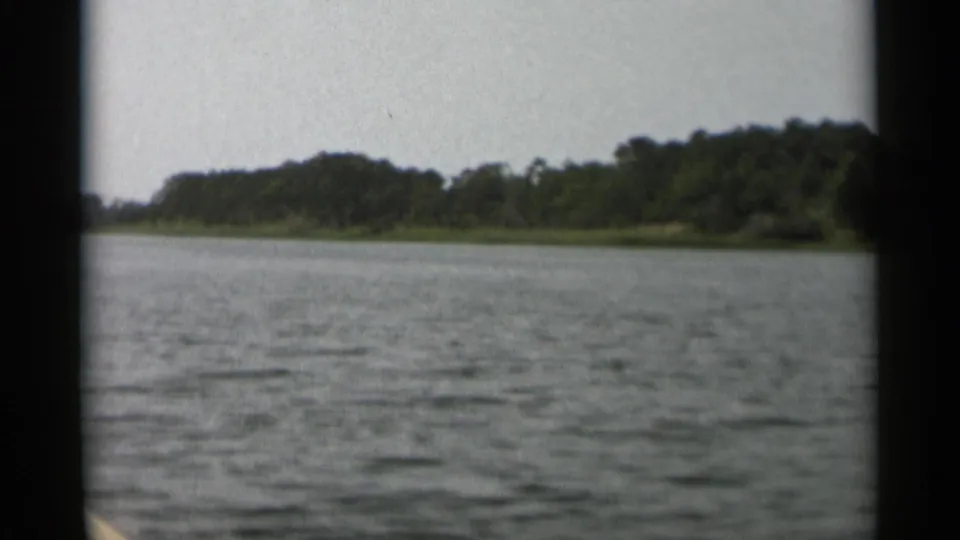 1956 Cape Cod frame showing rippled lake water and a wooded shoreline from the perspective of a small boat.