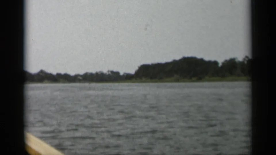 Boat-level 1956 Cape Cod view across rippled water toward low wooded land and a pale rail in the foreground.