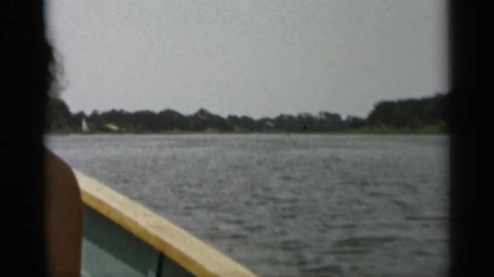 1956 Cape Cod boating frame with a partial passenger at left, the boat rail below, and distant shoreline across the water.