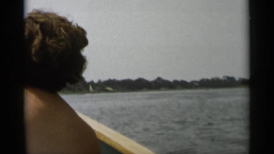 A curly-haired passenger seen from behind on a 1956 Cape Cod boat trip, facing the water and low wooded shore.