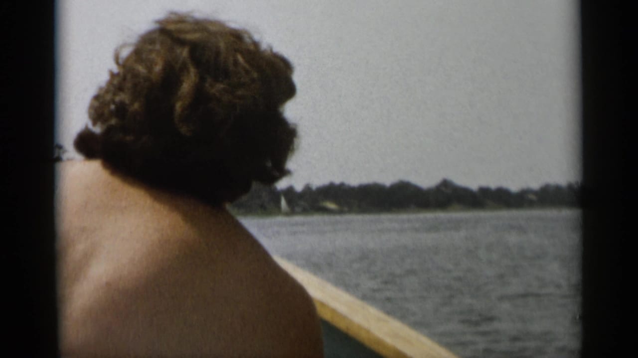 1956 Cape Cod home-movie frame showing a shirtless man from behind as a small boat moves across calm water near a wooded shore.