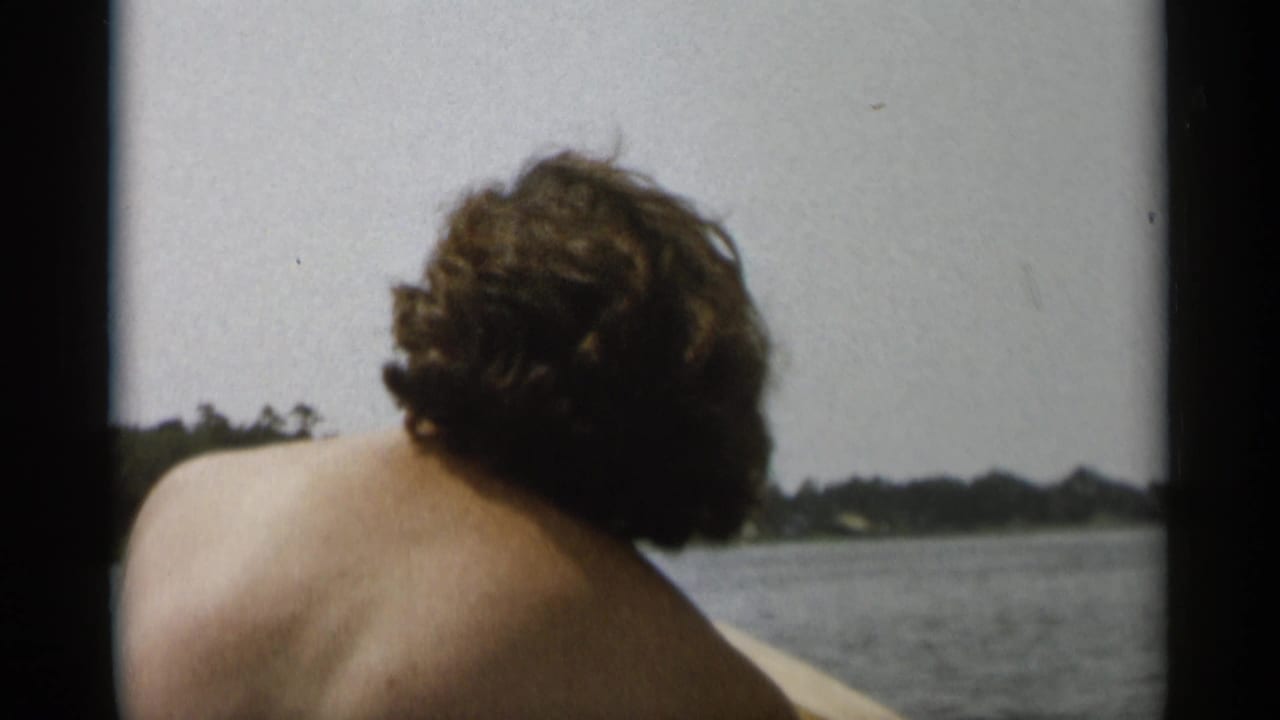 Cape Cod 1956 frame with a shirtless passenger viewed from behind, open water around the boat, and a low tree-lined shore in the distance.