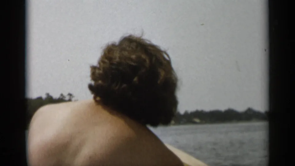 Cape Cod 1956 frame with a shirtless passenger viewed from behind, open water around the boat, and a low tree-lined shore in the distance.