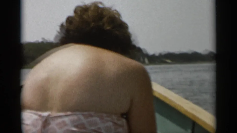 1956 Cape Cod boating frame showing the passenger leaning forward, with calm water, a wooden rail, and distant shoreline visible behind him.