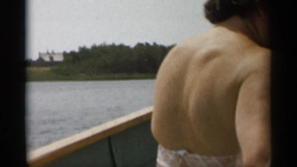 Cape Cod 1956 home-movie frame showing a seated passenger from behind beside calm water, trees, and a distant light-colored house.