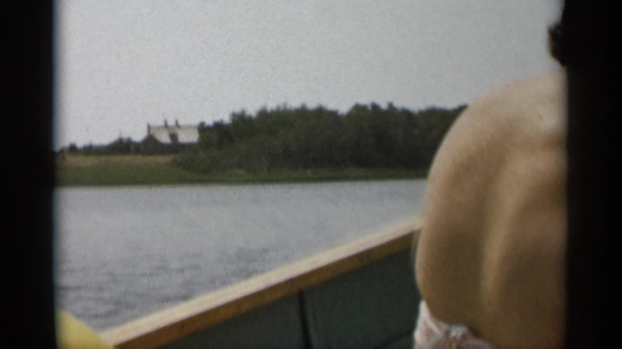 View from a boat across calm water with a passenger's bare back and a distant shoreline house.