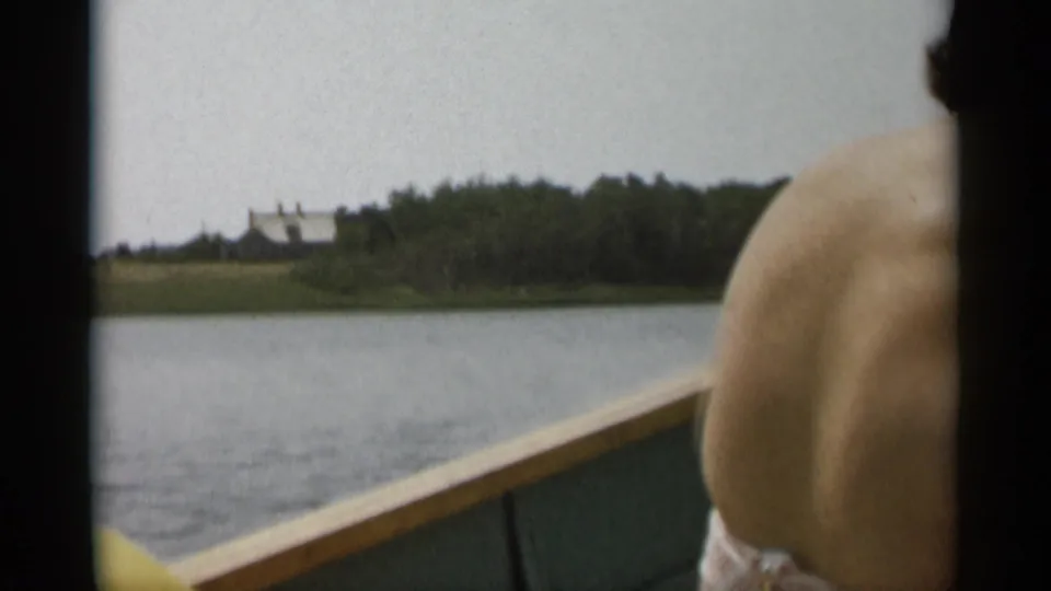 1956 Cape Cod 8mm frame from a boat, showing calm water, a wooded shore, a distant white house, and part of a passenger in the foreground.