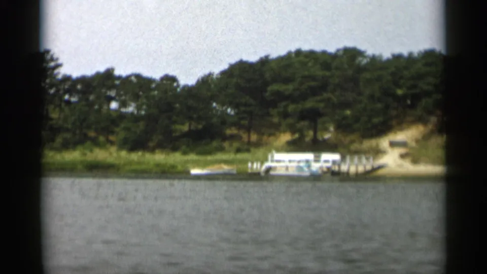 1956 home-movie frame of a Cape Cod waterfront with a dock, white boats, grassy bank, sandy rise, and trees behind it.