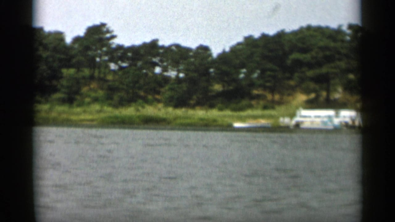 1956 Cape Cod 8mm frame showing bay water, dense trees, sandy shoreline, and small boats docked along the edge.