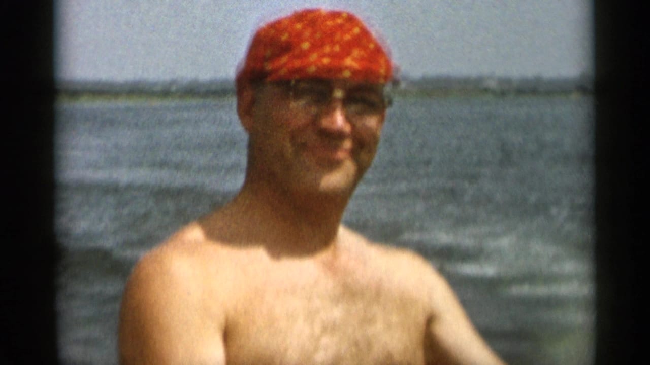 1956 Cape Cod home-movie frame of a shirtless adult man in glasses and a red patterned cap with bay water behind him.