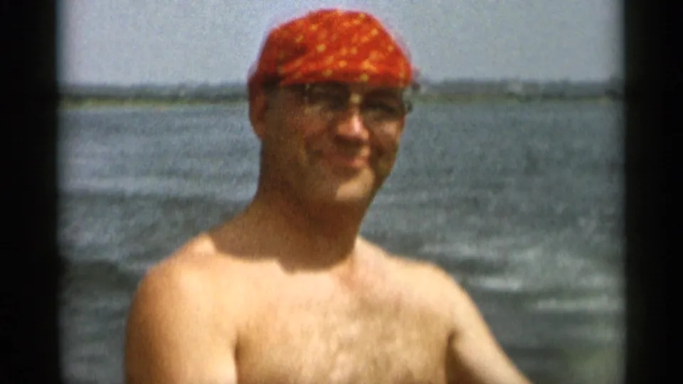 1956 Cape Cod home-movie frame of a shirtless adult man in glasses and a red patterned cap with bay water behind him.
